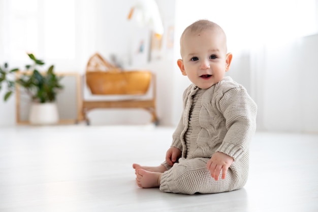 Bebezinho bonito em um terno de malha de lã aprendendo a sentar no chão em uma sala de estar brilhante bebê sorrindo brincando com o desenvolvimento inicial das crianças