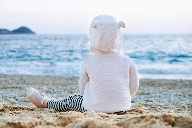 Bebezinho caucasiano sentado na praia olhando para o marVista de trás Camisola infantil bonita com orelhas de rato engraçadas passando tempo à beira-marRoupas de criança adoráveis