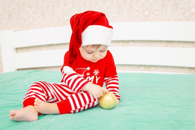 Bebezinho com chapéu de Papai Noel brincando com bola dourada de natal na cama ano novo e férias de inverno