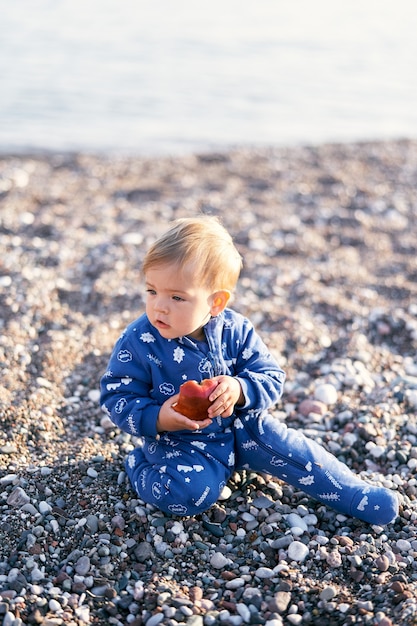 Bebezinho de macacão azul sentado em uma praia de seixos segurando uma maçã close-up