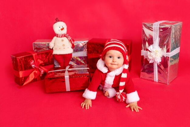 Bebezinho feliz com fantasia de Papai Noel sentado perto de presentes em vermelho isolado, conceito de ano novo