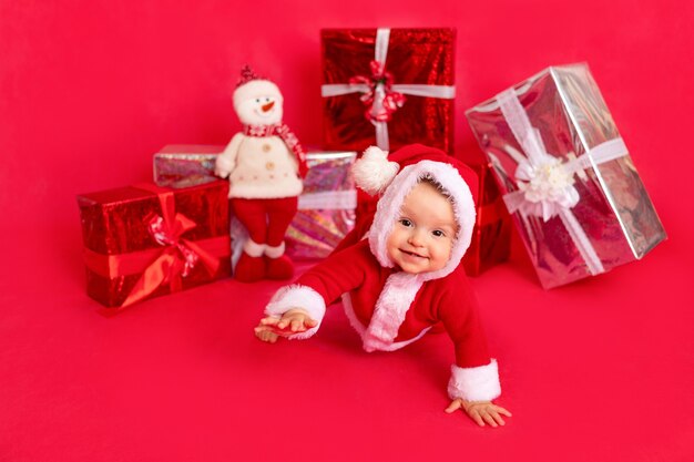 Bebezinho feliz de seis meses com fantasia de Papai Noel sentado perto de presentes no fotofone vermelho isolado, espaço para texto, ano novo e conceito de férias