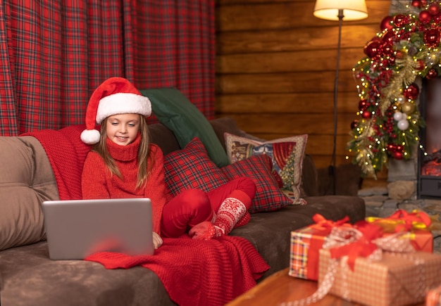 Foto bebezinho fofo com um chapéu de papai noel, sentado no sofá, olhando para um laptop com um sorriso antes do natal