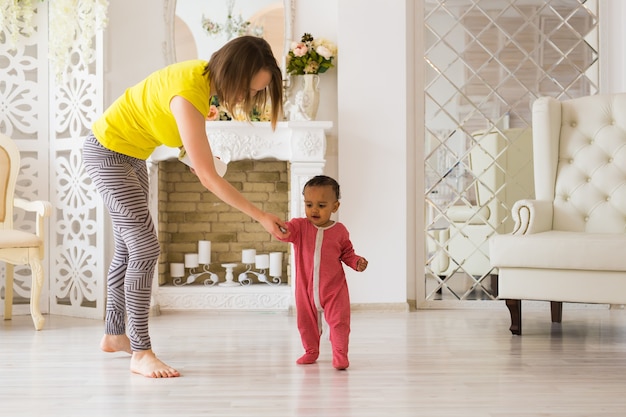 Foto bebezinho fofo de raça mista aprendendo a andar, a mãe segurando suas mãos
