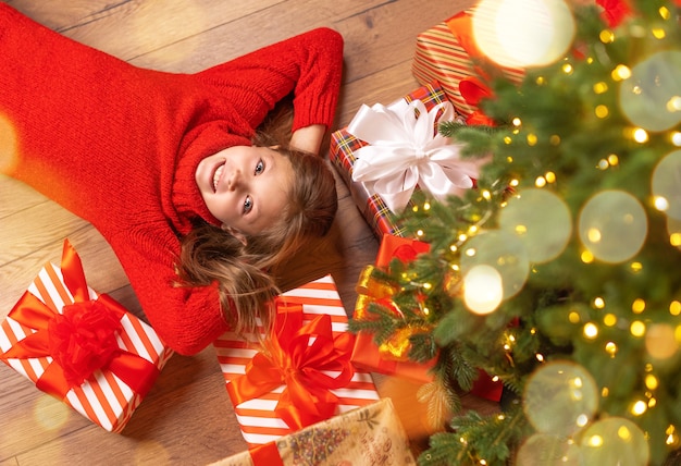 Bebezinho fofo está deitado no chão perto da árvore de Natal com presentes e olha para a câmera com um sorriso e alegria em casa