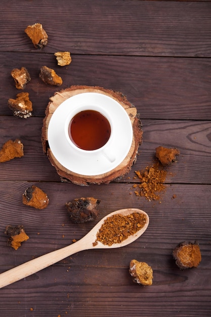 Bebida de café con hongos chaga en una taza blanca sobre un soporte de madera trozos dispersos de chaga y una cuchara con polvo de chaga sobre un fondo de madera vista superior vertical