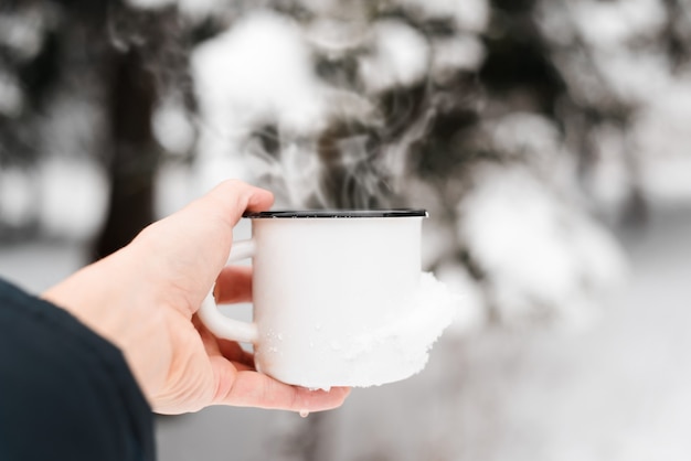 Bebida caliente en clima frío. Mano sosteniendo la taza de acero con bebida caliente al aire libre, primer plano. Vapor saliendo de la taza