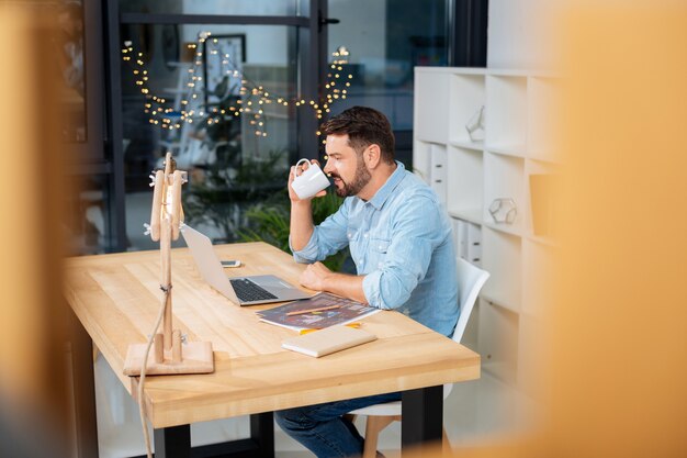 Bebida caliente. Hombre inteligente agradable agradable sosteniendo una taza y tomando un sorbo de té mientras trabaja en la computadora portátil