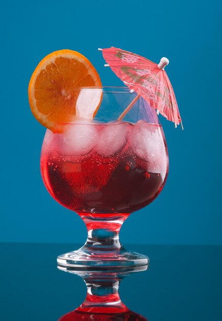 Bebida de cóctel rojo con cubitos de hielo sobre fondo azul.