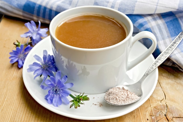 Bebida de chicória em um copo branco com uma flor e uma colher no pires, guardanapo sobre um fundo de tábuas de madeira