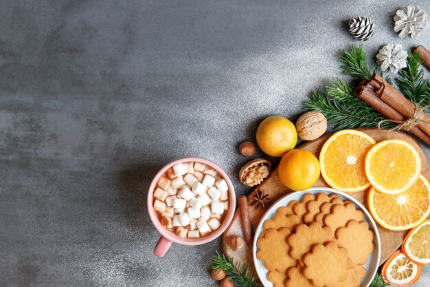 Bebida de inverno. Postura plana da caneca com chocolate quente ou cacau e pequenos pedaços de marshmallow, biscoito de gengibre, paus de canela, cones, raminhos de abeto, tangerina, nozes em fundo preto.