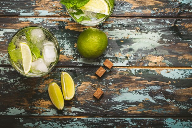 Bebida de verão em vidro e ingredientes para fazer Mojito com gelo, açúcar mascavo, fatias de limão, hortelã fresca em fundo rústico de madeira, espaço para texto. Coquetel refrescante para festa de verão e diversão