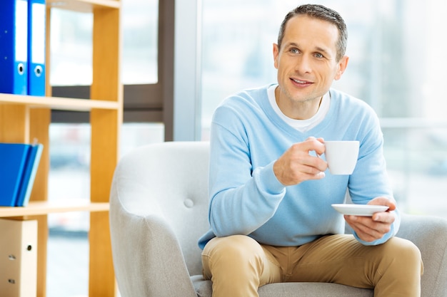 Bebida favorita. Agradable hombre positivo encantado agujereando una taza y bebiendo té mientras está sentado en el sillón en casa