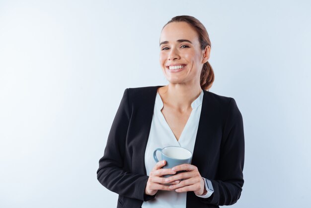 Bebida favorita. Alegre empresaria atractiva positiva sosteniendo una taza y disfrutando de su té mientras está de buen humor