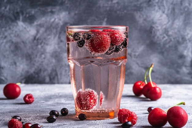 Bebida fresca de agua fría con gas con cereza, frambuesa y grosellas en vidrio rojo facetado sobre superficie de hormigón de piedra, bebida de dieta de verano, vista de ángulo