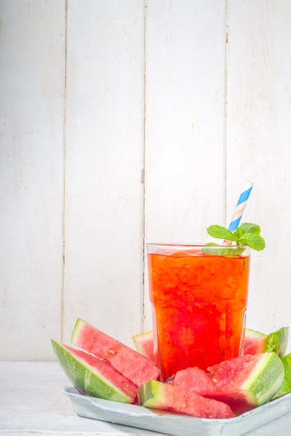 Bebida gelada gelada, mojito de batido de melancia ou suco, com fatias de melancia fresca, em uma pequena bandeja sobre uma mesa de madeira branca