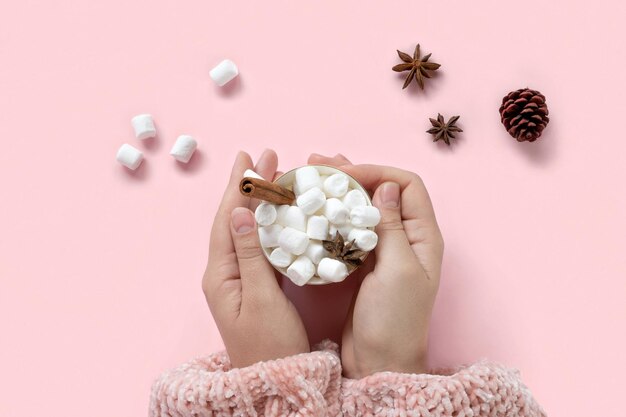 Bebida de invierno, taza blanca con malvaviscos en manos femeninas en suéter de Navidad tejido sobre fondo rosa. Vista superior, endecha plana. Año nuevo, comida tradicional navideña. Decoración festiva, celebración de Navidad.