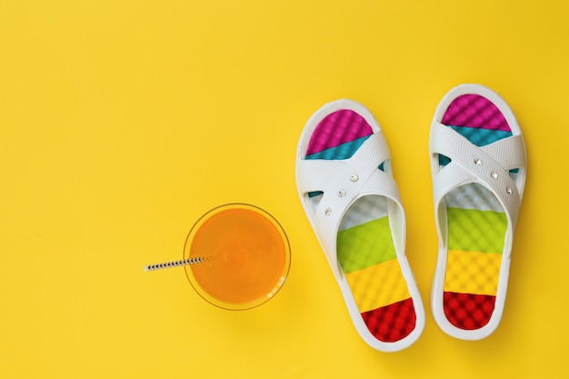 Bebida de naranja y pizarras con suelas de colores sobre fondo amarillo. El concepto de vacaciones de verano. Endecha plana. La vista desde arriba.