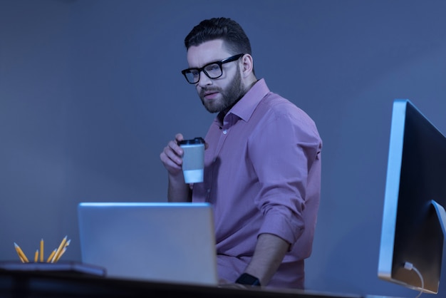 Bebida quente. homem sério bonito e agradável segurando uma xícara com café e olhando para a tela do laptop enquanto está sentado na mesa