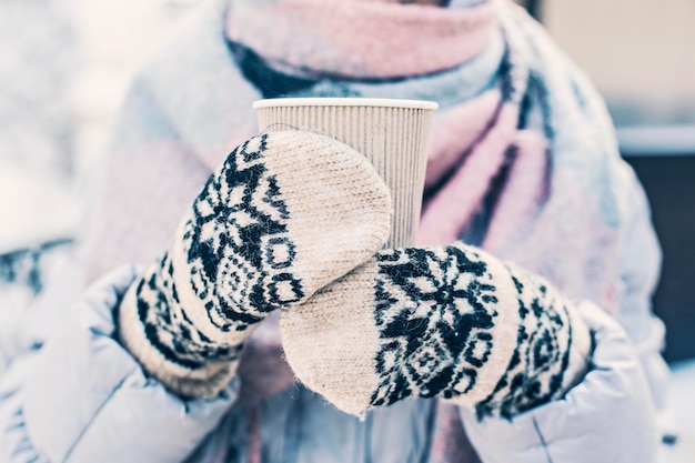 Bebida quente nas mãos da mulher usando luvas de malha quentes em dia frio de inverno nevado.