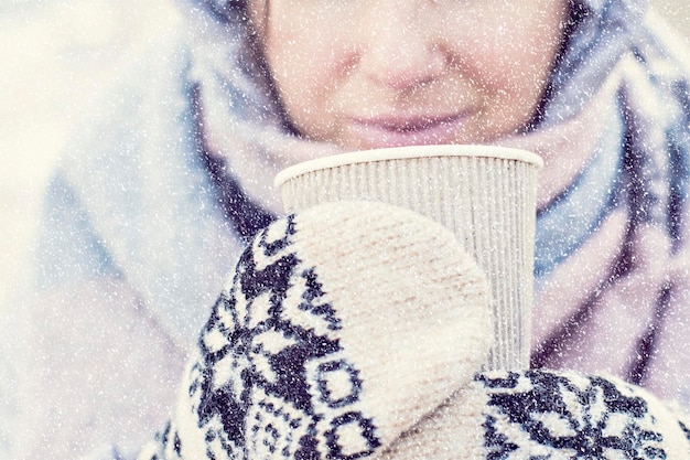 Bebida quente nas mãos da mulher usando luvas de malha quentes em dia frio de inverno nevado.