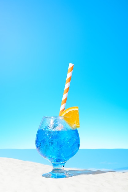 Bebida refrescante azul con hielo y una rodaja de naranja en un vaso en la costa arenosa
