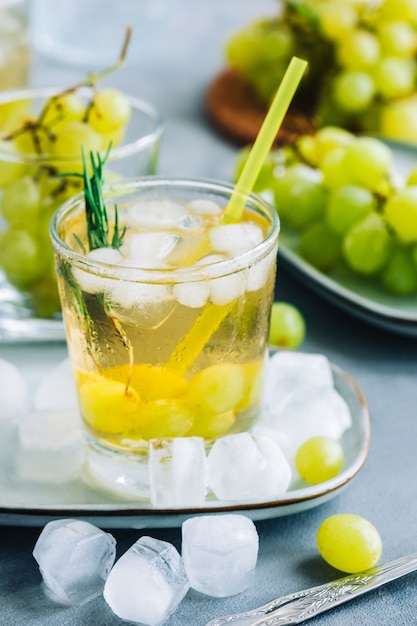 Bebida refrescante de verão em copo com cubos de gelo, uva verde e alecrim em cima da mesa.