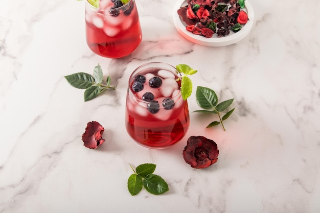 Bebida tónica de vitamina de hibisco de pétalos de rosa con hojas de menta de arándanos cubitos de hielo en vasos sobre una mesa de mármol té helado