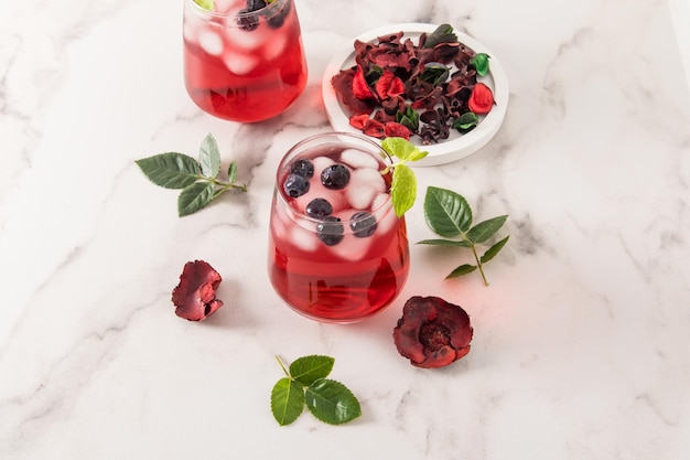 Bebida tónica de vitamina de hibisco de pétalos de rosa con hojas de menta de arándanos cubitos de hielo en vasos sobre una mesa de mármol té helado