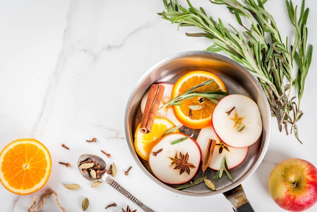 Bebida tradicional do inverno e do natal, ingredientes para a bebida quente quente com vinho cítrico, maçã e especiarias na caçarola de alumínio na mesa de mármore branca. copie o espaço vista superior