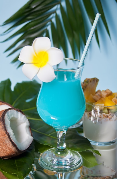 Bebida tropical exótica do cocktail de Curaçau azul em um copo com flor de frangipani do Plumeria, folha de palmeira, coco fresco no fundo.