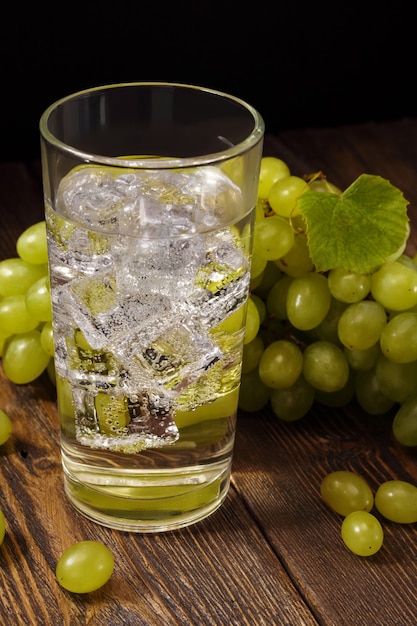 Bebida de uva fresca con cubitos de hielo en vaso y racimo de uva verde dulce en mesa de madera.