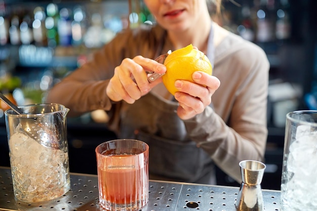 bebidas alcohólicas, gente y concepto de lujo - mujer barman con vaso y pelador quitando la cáscara de naranja y preparando cócteles en el bar