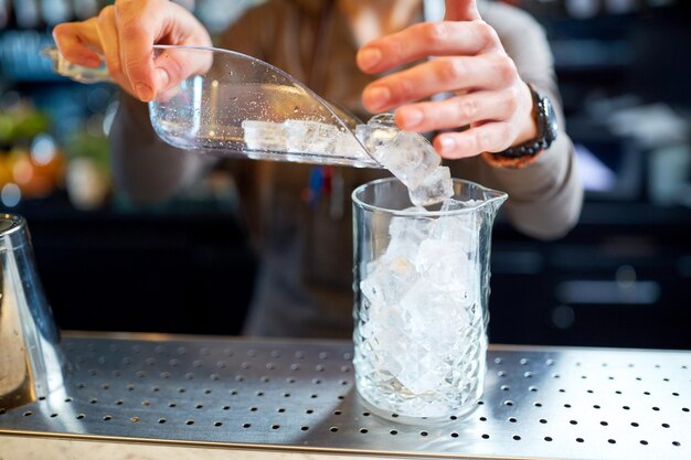 Foto bebidas alcoólicas, pessoas e conceito de luxo - barman com pá derramando gelo em jarra de vidro e preparando coquetel no balcão do bar