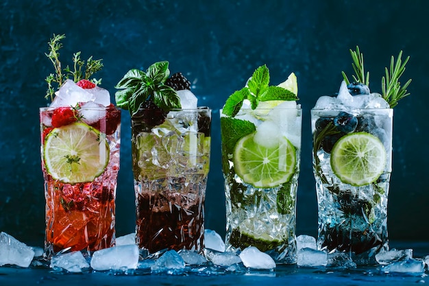 Bebidas de coquetéis frios de verão bebida longa alcoólica clássica e mojito ou mocktail em highballs com mirtilos amoras framboesas ervas de limão e gelo sobre fundo azul