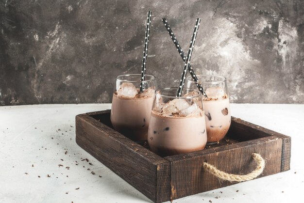 Bebidas de verão. Cacau gelado gelado de chocolate. Com uma colher de sorvete de chocolate, chocolate em pó e gelo. Em copos, com tubos para beber. Bandeja de madeira mesa de concreto branco.