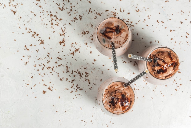 Bebidas de verão. cacau gelado gelado de chocolate. com uma colher de sorvete de chocolate, chocolate em pó e gelo. em copos, com tubos para beber. mesa de concreto branca. vista do topo