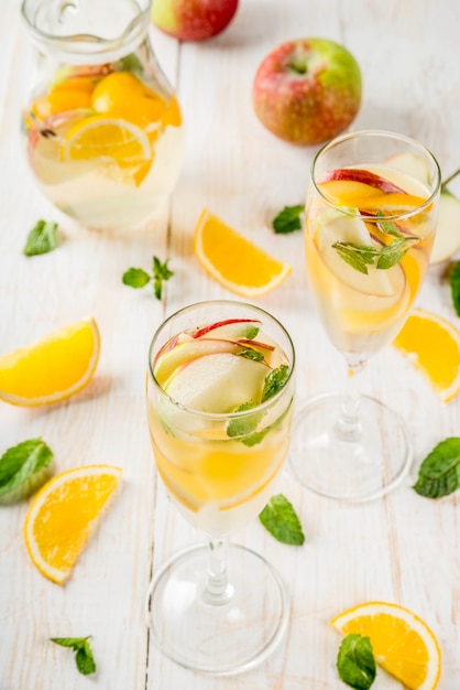 Bebidas e coquetéis. Sangria de outono branca com maçãs, laranja, hortelã e vinho branco. Em copos de champanhe, em uma jarra, em uma mesa de madeira branca.