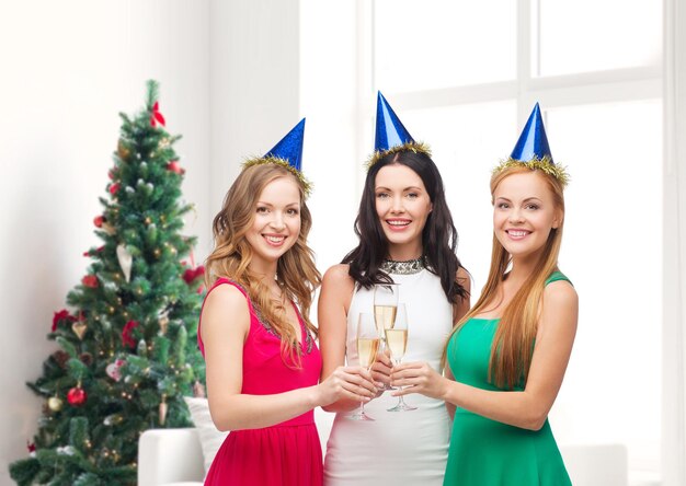 bebidas, feriados, pessoas e conceito de celebração - mulheres sorridentes em chapéus de festa com copos de vinho espumante sobre a sala de estar e o fundo da árvore de natal