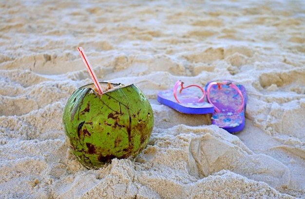 Bebidas frescas de coco joven con un par de sandalias borrosas en la playa de arena