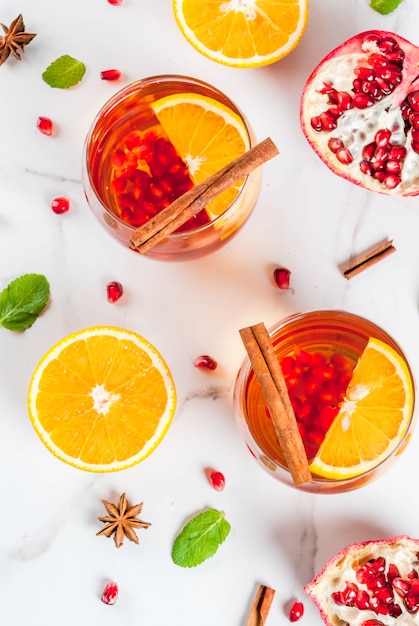 Bebidas de otoño e invierno. Cóctel cálido y refrescante con granada, naranjas, canela, especias y menta. Sobre una mesa de mármol blanco. vista superior