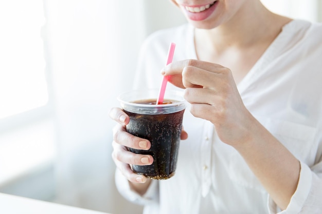 bebidas, pessoas e conceito de estilo de vida - close-up de mulher feliz bebendo coca cola de copo de plástico com palha em casa