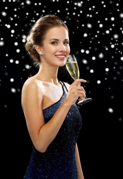 bebidas, vacaciones de invierno, navidad y concepto de la gente - mujer sonriente vestida de noche con una copa de vino espumoso sobre fondo negro nevado