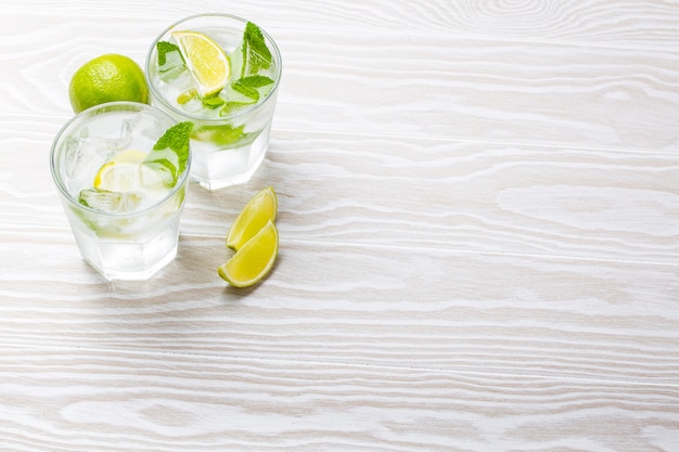 Bebidas de verano con hielo, rodajas de limón, menta fresca en vasos sobre fondo rústico de madera blanca, espacio para texto. Limonadas / mojito refrescantes frías ideales para fiestas de verano y diversión, espacio de copia, vista de ángulo