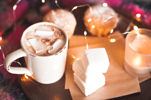 Becher mit Kaffee und Marshmallow mit Kuchen und Kerze bleiben auf Holztablett über Weihnachtsbeleuchtung Hintergrund Guten Morgen Selektiver Fokus