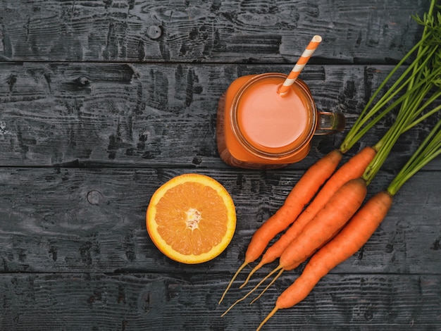 Becher mit Karotten-Smoothie, Ingwerwurzel und Orange auf rustikalem dunklem Holztisch.