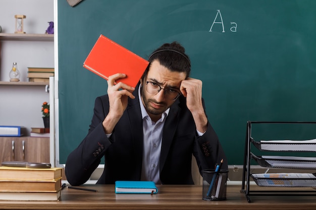 Bedauert, dass der männliche Lehrer mit gesenktem Kopf eine Brille trägt und ein Buch am Tisch mit Schulwerkzeugen im Klassenzimmer sitzt