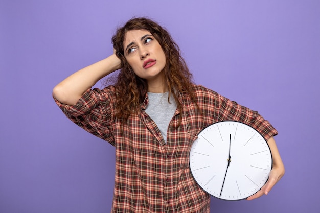 Bedauert, die Hand auf den Kopf zu legen, eine junge Putzfrau, die eine Wanduhr isoliert auf einer lila Wand hält?