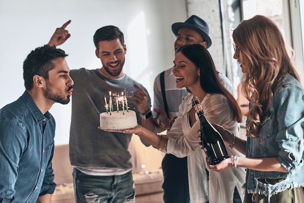 Beeilen Sie sich, um etwas zu wünschen. Glücklicher junger Mann, der Kerzen bläst, während er Geburtstag unter Freunden feiert