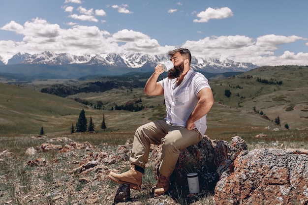 Beeindruckende und ruhige Berglandschaft. Ein erwachsener Mann mit Bart in einem weißen Hemd sitzt auf einem Stein auf einem grünen Rasen und trinkt heißen Tee. Unter dem Kopfsteinpflaster befindet sich eine weiße Thermoskanne.